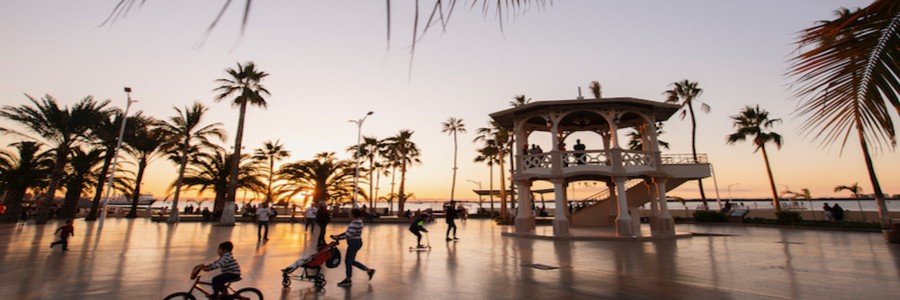 A lively sunset scene along the Malecón in La Paz, Mexico, with palm trees, people walking and biking, and a seaside pavilion overlooking the ocean under a glowing sky