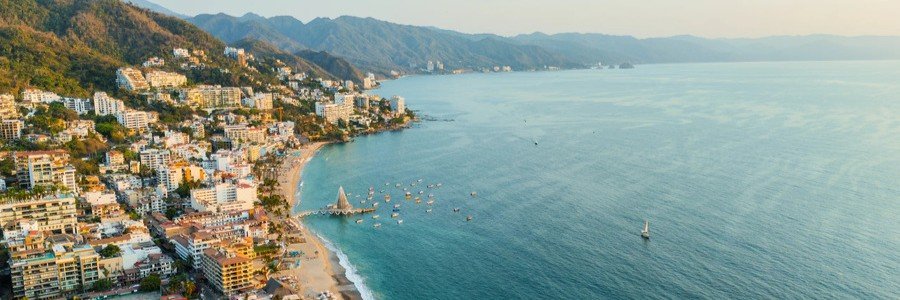 A scenic aerial view of La Paz, Mexico showing the coastline lined with colorful buildings, a sandy beach, and calm blue waters stretching across the bay, with mountains rising in the background under a clear sky