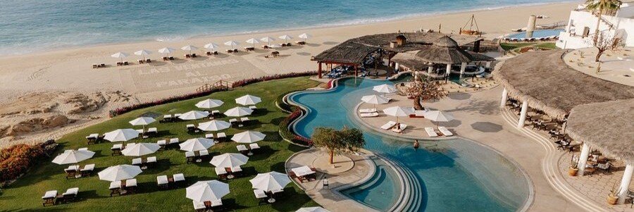 Aerial view of Las Ventanas al Paraiso in Cabo San Lucas featuring a curved infinity pool, white loungers and umbrellas on green lawns, beachfront cabanas, and calm blue ocean along a sandy shoreline