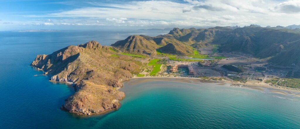 loreto-baja-california-coastal-aerial-mountain-bay | Beach Travel Destinations A wide aerial view of Loreto, Baja California Sur, showing a curved bay with calm turquoise water, rugged desert mountains, and a coastal resort area along the shoreline under a partly cloudy sky