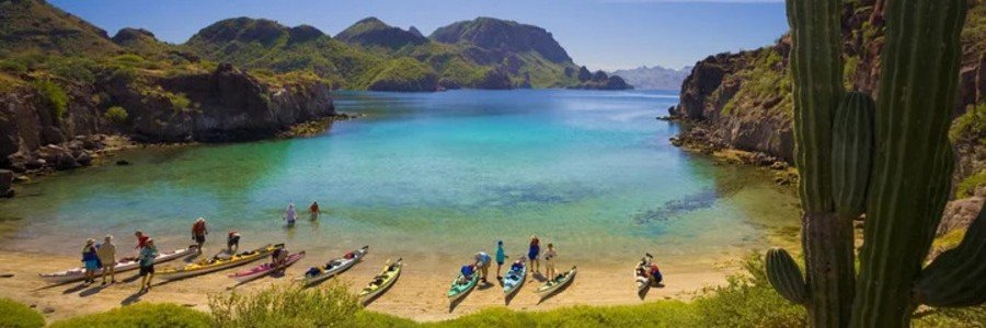 loreto-best-beaches-kayaking-cove | Beach Travel Destinations Colorful kayaks lined along a sandy beach in a turquoise cove in Loreto, Mexico, with mountains and cactus surrounding the calm water