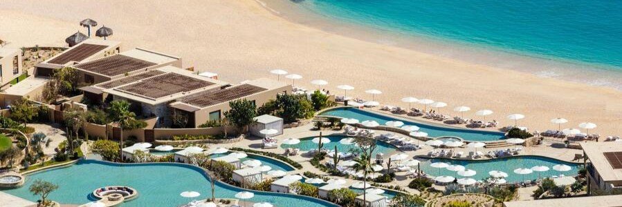 Aerial view of Montage Los Cabos with multiple curved infinity pools, white umbrellas and lounge chairs, modern villas, and a calm turquoise bay along a sandy beach