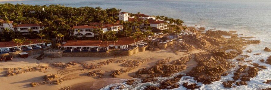 Aerial view of One and Only Palmilla in Cabo San Lucas with red-tiled roofs, palm trees, sandy beach, and rocky shoreline along the Sea of Cortez at golden hour