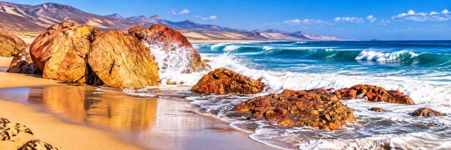 Panoramic view of Piedra Playa near El Cotillo, with rust-colored volcanic rocks, powerful Atlantic waves, golden sand, and rugged coastal scenery.
