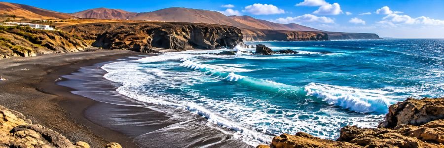 Panoramic view of Playa de Ajuy in Fuerteventura, featuring black volcanic sand, turquoise Atlantic waves, rugged coastal cliffs, and mountains beneath a blue sky.