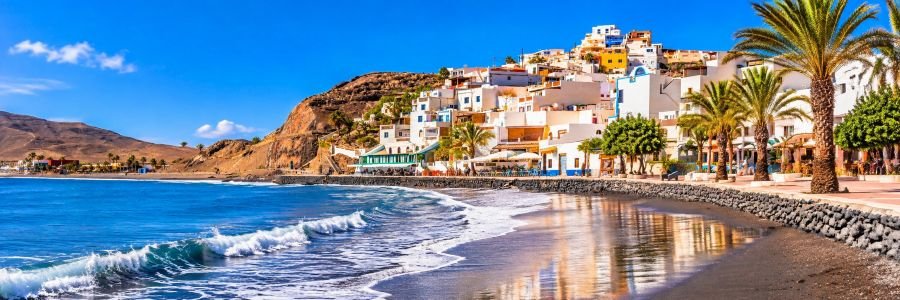 Panoramic view of Playa de Gran Tarajal in Fuerteventura, featuring dark volcanic sand, blue Atlantic waters, whitewashed hillside buildings, and a palm-lined seaside promenade.