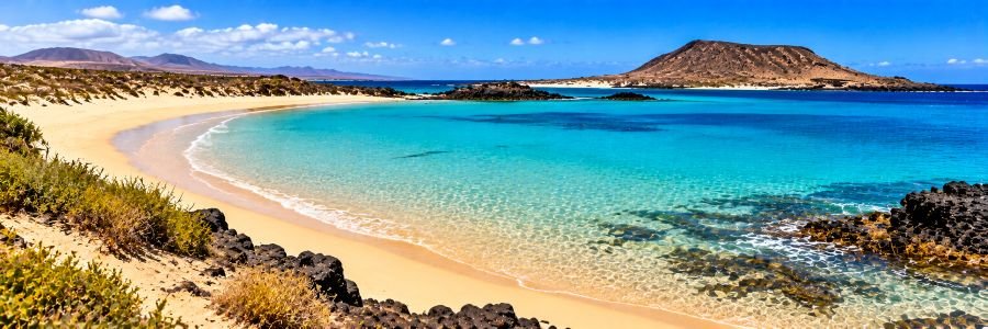 Panoramic view of Playa de La Concha on Lobos Island, featuring a crescent-shaped sandy beach, crystal-clear turquoise water, volcanic rocks, and distant mountains.