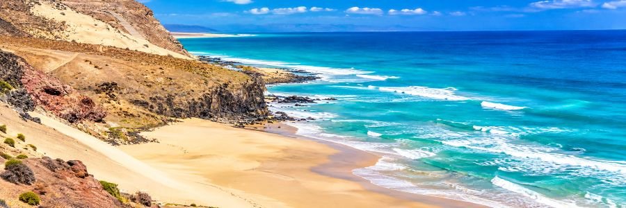 Panoramic view of Playa de Mal Nombre in southern Fuerteventura, featuring golden sand, rugged coastal cliffs, turquoise Atlantic waters, and gentle waves along a secluded shoreline.