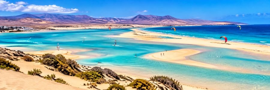 Panoramic view of Playa de Sotavento in Fuerteventura, with turquoise tidal lagoons, golden sandbars, colorful kitesurfers, and volcanic mountains.