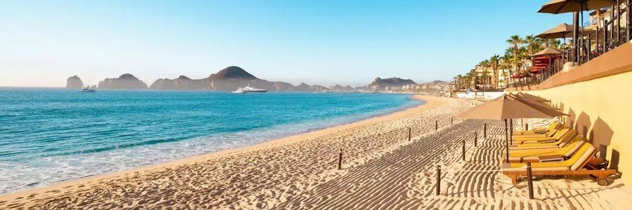 Sunny beachfront at Playa Palmilla Los Cabos with golden sand, turquoise water, lounge chairs, and coastal mountain views.
