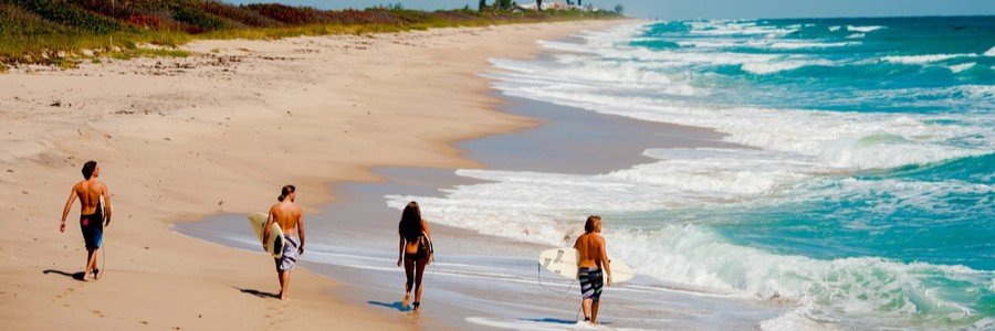 Four young surfers carrying surfboards walk along a wide sandy shoreline as vibrant turquoise Atlantic waves crash onto the beach.