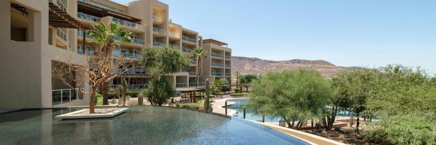Puerta Cortez Hotel in La Paz featuring a modern resort pool surrounded by desert landscaping, palm trees, and multi-level buildings with balconies overlooking the Baja desert hills.