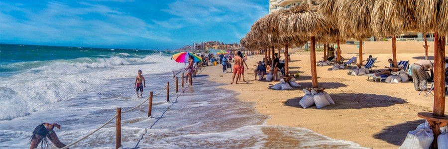 Busy beach scene at Puerto Peñasco in Mexico with waves rolling onto the sand, shaded palapas, beach chairs, and visitors enjoying the sunny Sea of Cortez coastline.