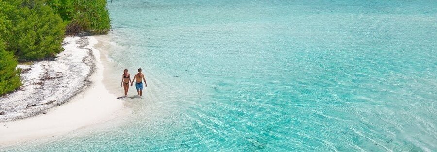 Couple walking along a white sand beach in Raivavae with clear turquoise water and lush tropical greenery in the Austral Islands