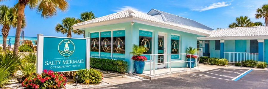 The Salty Mermaid Oceanfront Hotel in New Smyrna Beach, featuring a turquoise exterior, tropical landscaping, and colorful flowers near the ocean.