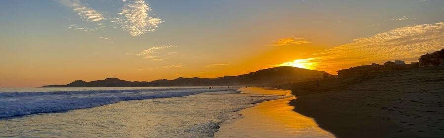 Golden sunset over the beach in San José del Cabo with glowing shoreline reflections, gentle ocean waves, and Baja mountain silhouettes.