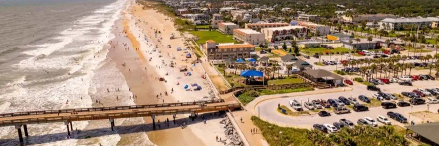 Aerial view of a sandy beach and pier extending into the ocean, with a town and parking area inland and people enjoying the shore.