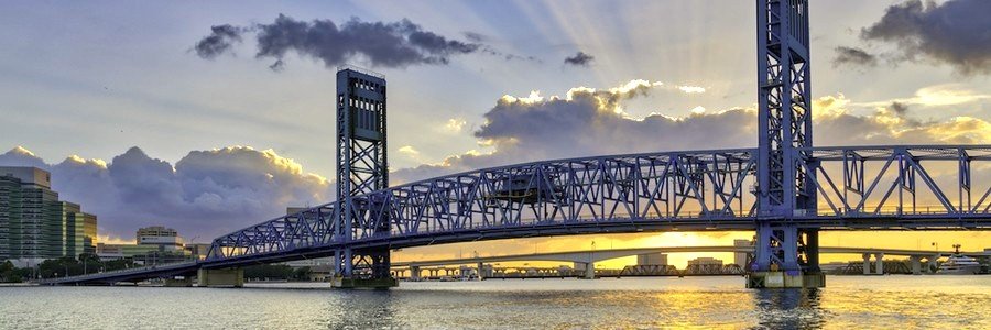 St Johns River flowing beneath Jacksonville's iconic blue bridge at sunset with golden reflections across the water.