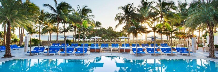 Oceanfront swimming pool at The St. Regis Bal Harbour Resort with rows of bright blue lounge chairs and palm trees at sunrise.