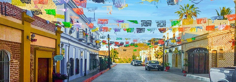 Colorful street in Todos Santos Mexico with papel picado banners, local shops, colonial buildings, and palm trees under blue sky