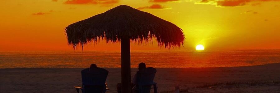 Sunset beach in Todos Santos Mexico with palm palapa umbrella, ocean horizon, and silhouettes watching the sun set over the Pacific