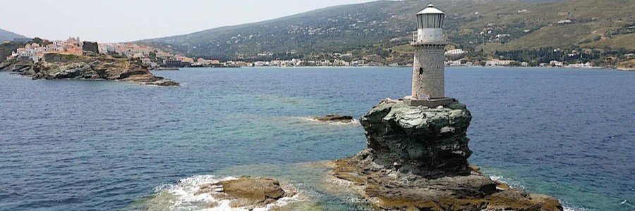 Tourlitis Lighthouse standing on a rocky sea stack near Chora on Andros Island, Greece, with the coastline and village in the background.