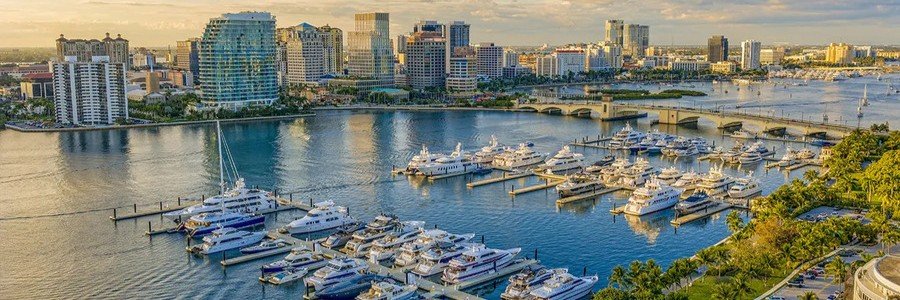 High-angle aerial view of luxury mega yachts docked at the Town of Palm Beach Marina, with the Royal Park Bridge and downtown West Palm Beach skyscrapers in the background.