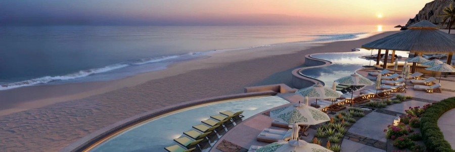 Sunset view at Waldorf Astoria Los Cabos Pedregal with an infinity pool overlooking the beach, lounge chairs and umbrellas on a terrace, and soft waves along the sandy shoreline under a colorful sky