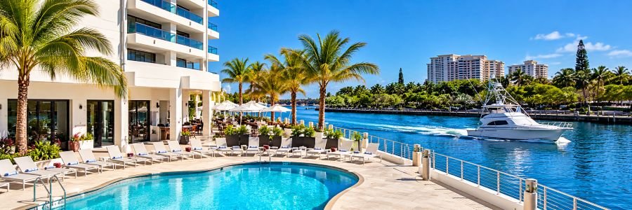 Luxury waterfront resort with a turquoise swimming pool, palm trees, lounge chairs, and a white yacht cruising along the marina.