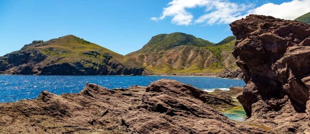 Rocky shoreline of Saba with rugged cliffs, blue ocean waters, and mountainous terrain under a bright sky.