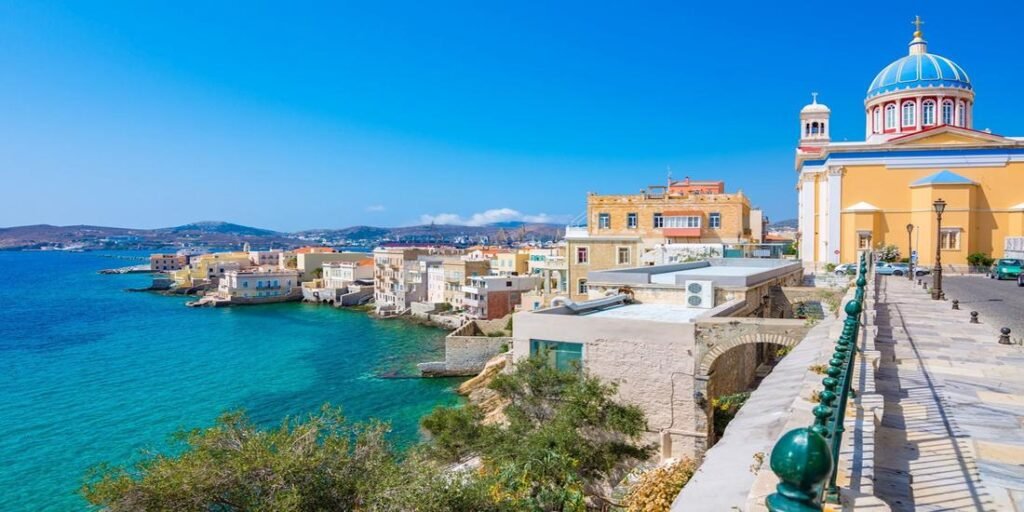 Most Beautiful Syros, Greece, with colorful neoclassical houses, a blue-domed church, and turquoise Aegean waters along the coast.