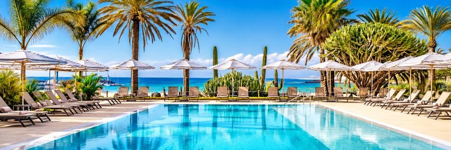 Sparkling turquoise swimming pool surrounded by striped umbrellas, comfortable lounge chairs, tall palm trees, and beautiful ocean views.