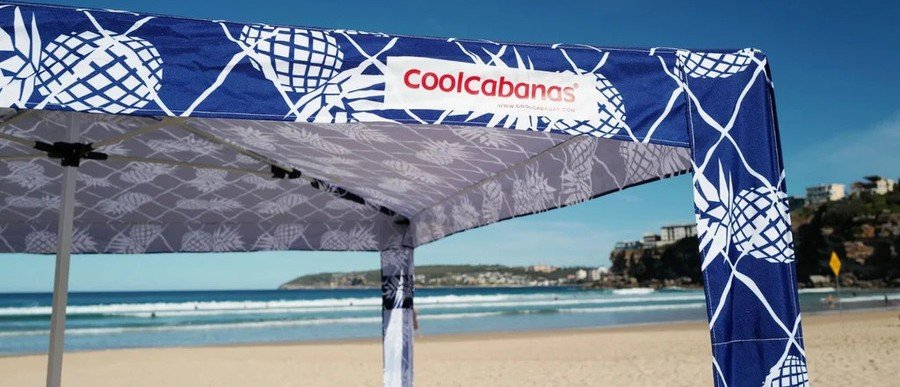 Blue-and-white pineapple-print CoolCabana 5 providing open-sided shade on a sandy beach beside the ocean.
