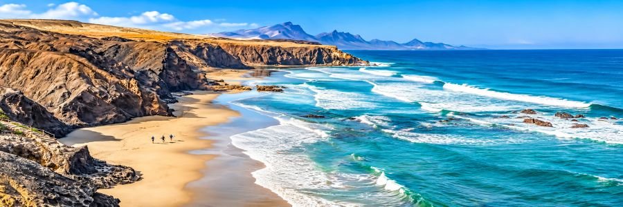 Panoramic view of golden sandy beaches, rugged coastal cliffs, turquoise Atlantic waters, and distant mountains in Fuerteventura.
