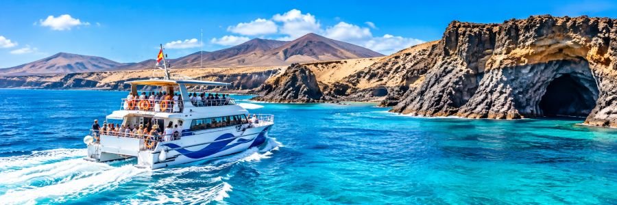Sightseeing catamaran cruising through turquoise Atlantic waters beside dramatic volcanic cliffs and sea caves in Fuerteventura.