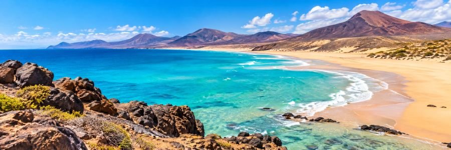 Panoramic Fuerteventura coastal landscape featuring turquoise Atlantic waters, golden sandy beaches, rugged volcanic mountains, and a bright blue sky in the Canary Islands.