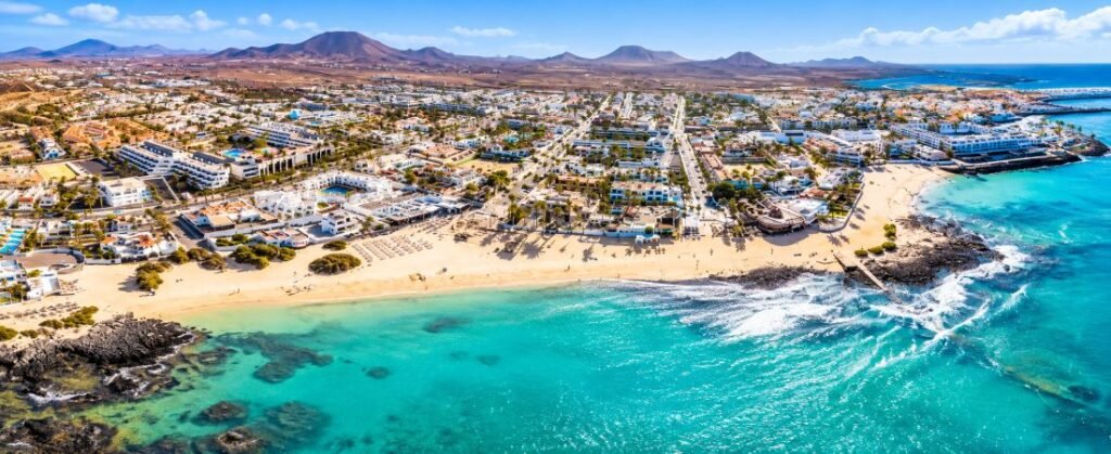 Panoramic aerial view of a Fuerteventura coastal resort, featuring turquoise Atlantic waters, golden sandy beaches, whitewashed buildings, and volcanic mountains.