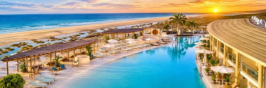 Expansive turquoise swimming pool surrounded by white lounge chairs, shaded pergolas, palm trees, and a sandy beach overlooking the ocean at sunset.