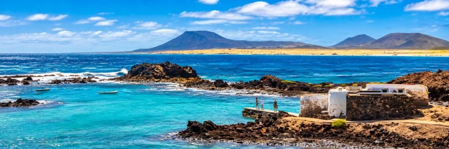Turquoise waters, black volcanic rocks, a white coastal building, and distant volcanic mountains at Isla de Lobos near Fuerteventura.