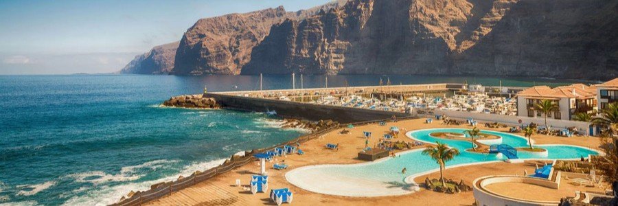 Towering dark volcanic basalt cliffs of Los Gigantes rising above a coastal resort pool and boat marina in Tenerife.