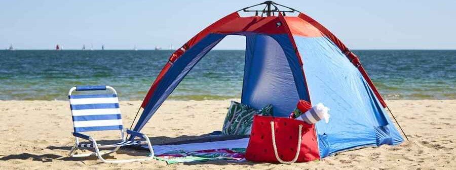 Red and blue Oileus XL Beach Tent set up beside a striped chair and beach bags near the ocean.