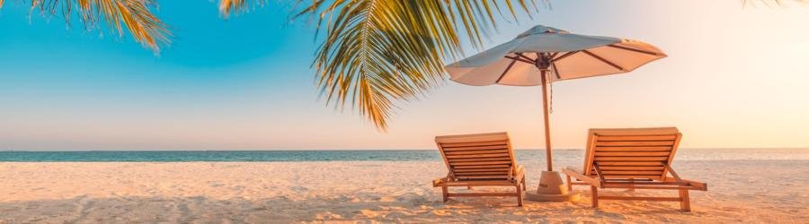 Wooden lounge chairs under a white beach umbrella at sunset with palm leaves overhead.