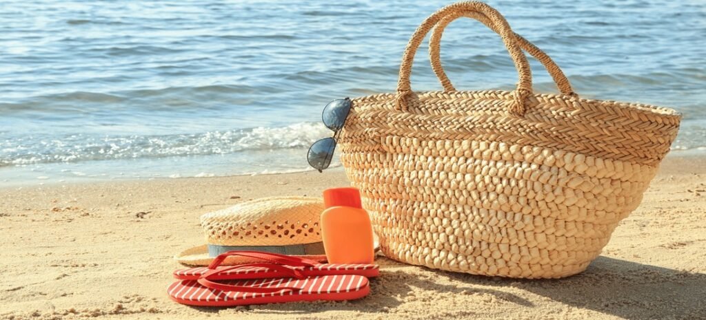 Straw beach bag with sunglasses, sunscreen bottle, sun hat, and red striped flip-flops on sandy shore with gentle waves in the background.