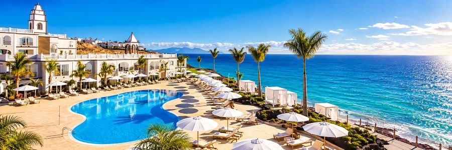 Curved blue swimming pool surrounded by white umbrellas, palm trees, elegant resort buildings, and sparkling ocean views.