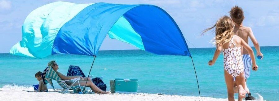 Blue Shibumi Shade Classic floating in the ocean breeze while a family relaxes and children run along the beach.