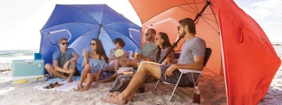 Families and friends relaxing beneath blue and orange Sport-Brella Premiere XL umbrella shelters on a sandy beach.