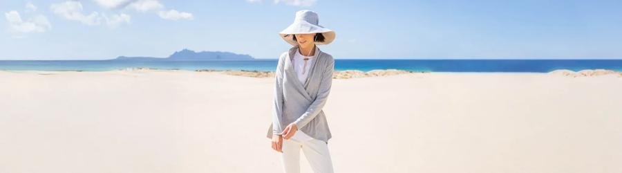 Woman wearing a wide-brimmed sun hat and light clothing on a bright sandy beach.
