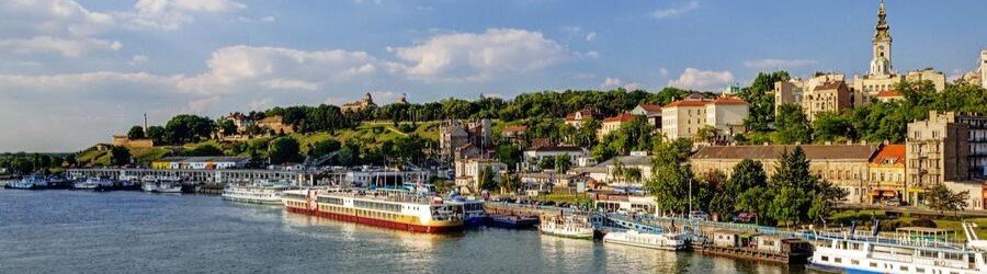 River cruise ships docked along the Danube River in Belgrade, Serbia, with the historic city skyline rising above the waterfront.