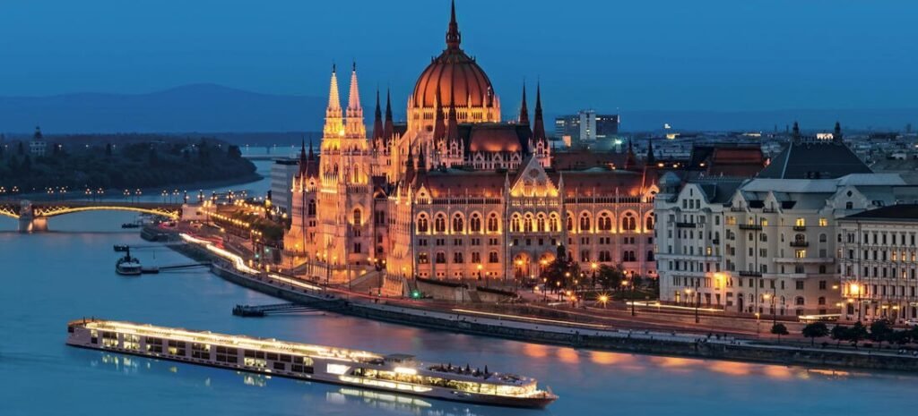 A luxury river cruise ship sailing past the illuminated Hungarian Parliament Building on the Danube River at night in Budapest.