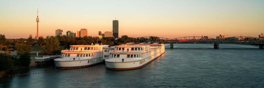 Two white luxury river cruise ships docked along the Danube River with the modern city skyline of Vienna Austria at sunset.